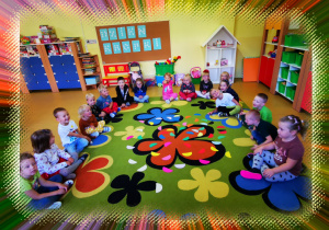 Grupa uśmiechniętych dzieci siedzi na dywanie w kolorowe kwiaty, na którym leżą papierowe półkola.
